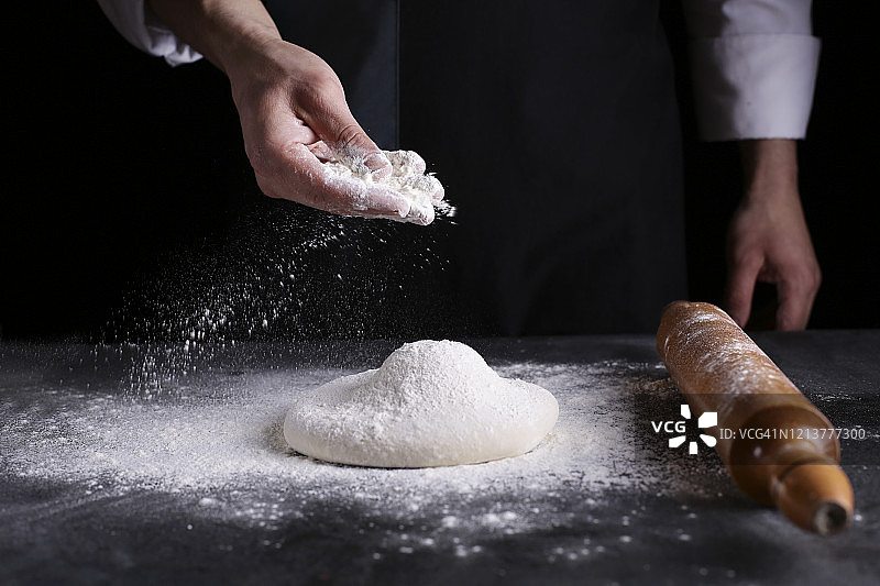 厨师用手将面粉粉倒入生面团上，用筛子在黑色的背景上，烹饪过程图片素材