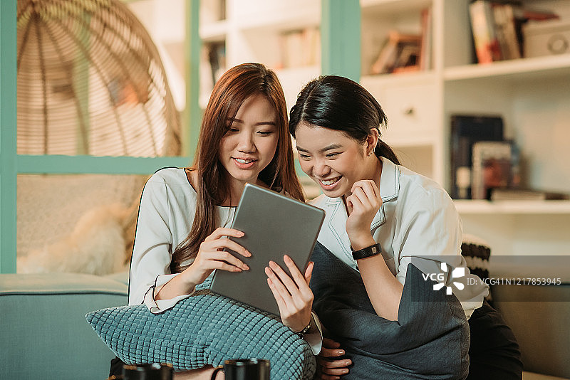 两位亚洲华人女商人微笑着坐在沙发上聊天，在室内办公休闲区的茶点休息时间用着平板电脑图片素材