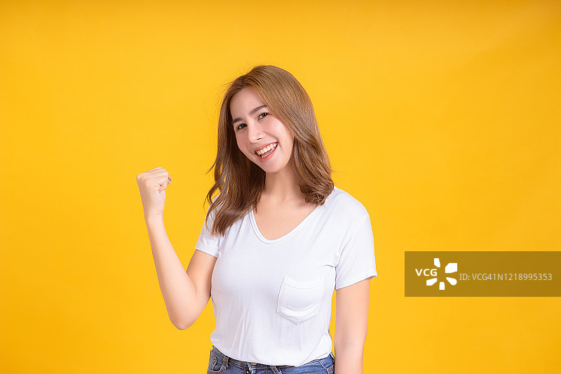 肖像快乐的年轻亚洲妇女微笑举起拳头幸运积极的情绪在白色t恤，黄色背景孤立的工作室拍摄和复制空间。图片素材