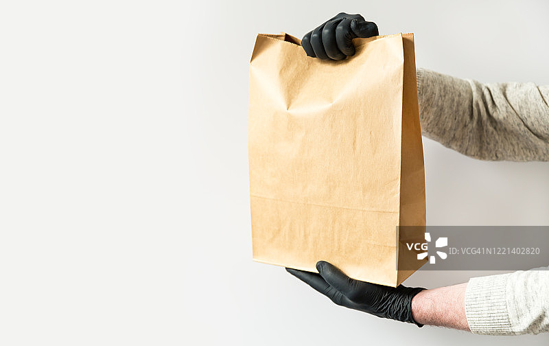 一个男人的手在黑色手套拿着纸袋的特写。白色背景。网上订餐，隔离期间非接触配送。图片素材