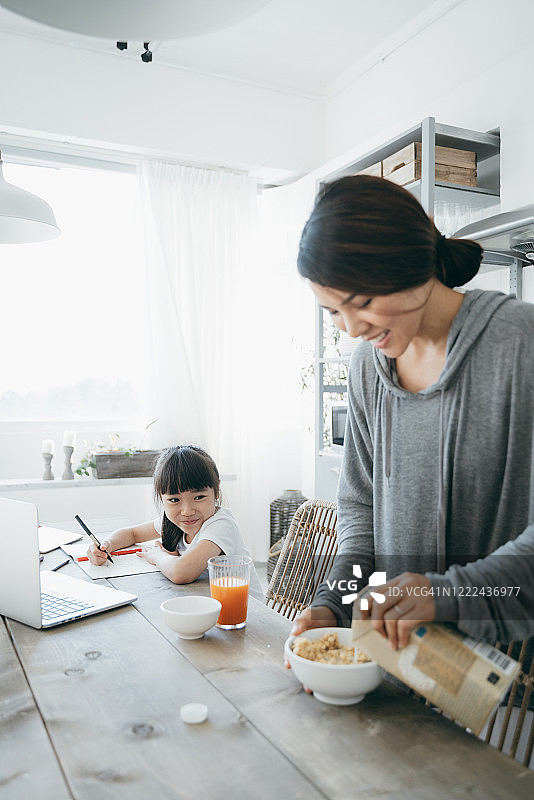 年轻的亚洲母亲准备健康的早餐，而小女儿正在家里学习。她用一台笔记本电脑在网上上课，在家写笔记图片素材