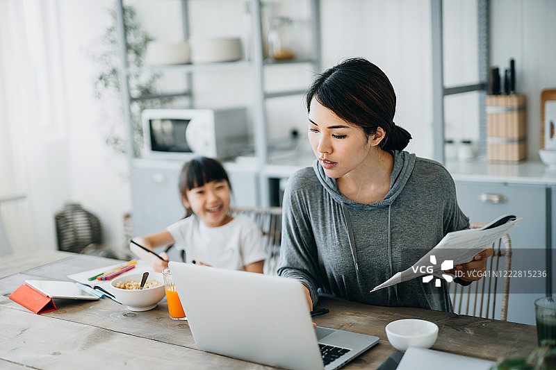 年轻的亚洲母亲在家里用笔记本电脑工作，而小女儿在家里学习。她用平板电脑上在线课程，在家做作业图片素材