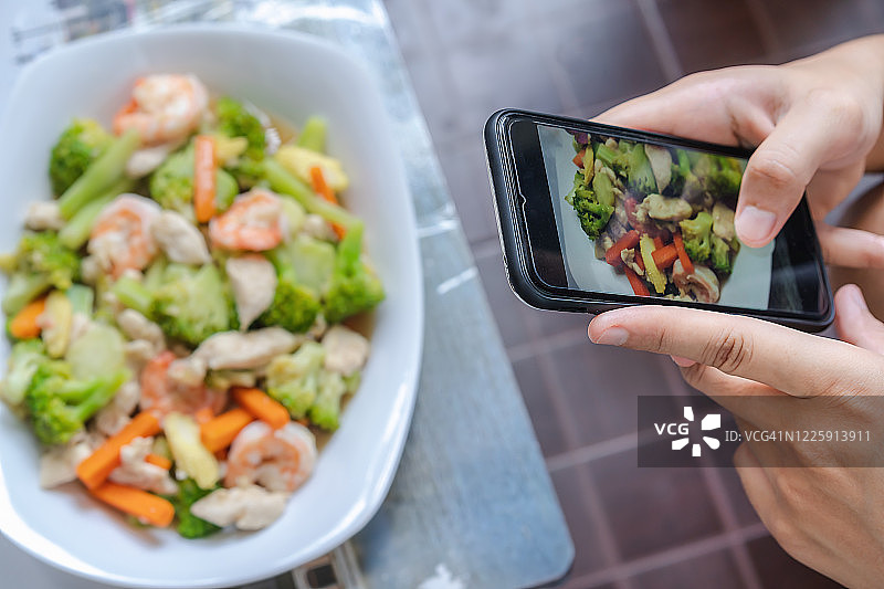 智能手机屏幕的特写用年轻女子的手指给食物拍照，模糊了混有虾和鸡的蔬菜。为社交网络拍摄午餐或晚餐照片。图片素材