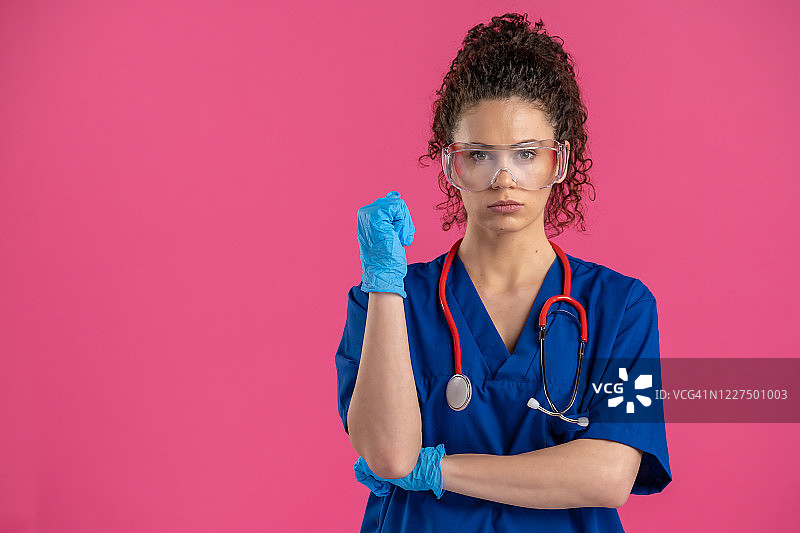 年轻的女医生戴着保护眼镜看着照相机，粉红色的背景图片素材
