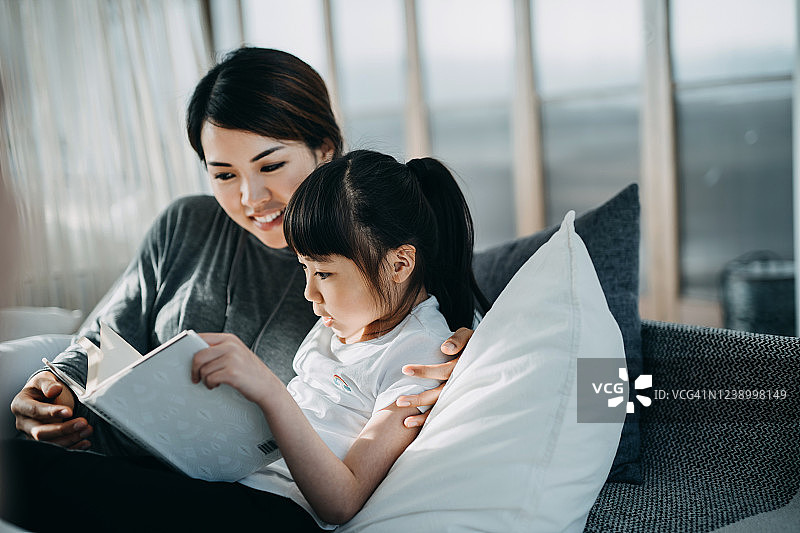 年轻的亚洲母亲在床上为小女儿读书，她们在家里享受家庭团聚的时光。美丽的阳光透过窗户照在他们身上图片素材