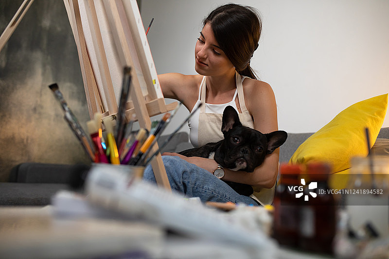 一个美丽的年轻女子在她的家里绘画，一条狗躺在她的腿上图片素材