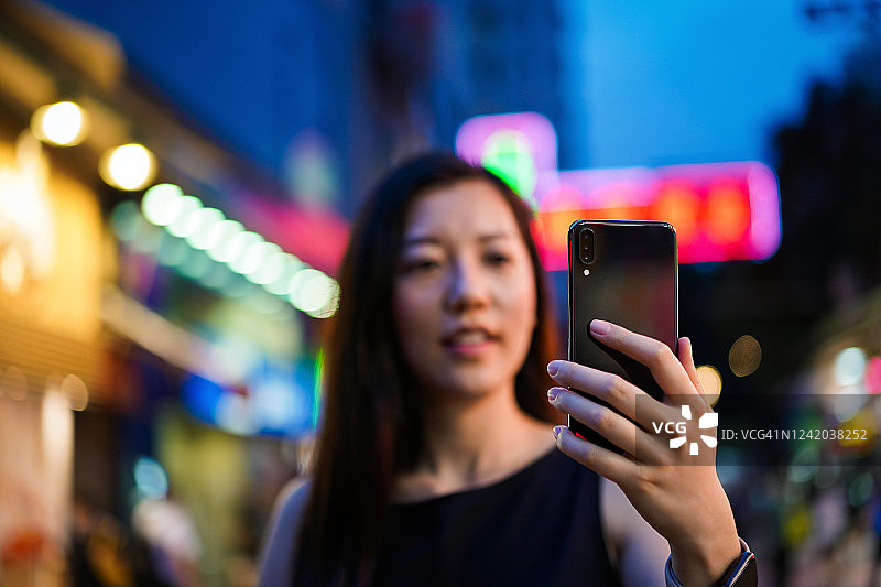 一名年轻的亚洲女性在繁忙的市中心街道上使用智能手机，站在五颜六色的霓虹灯招牌前图片素材