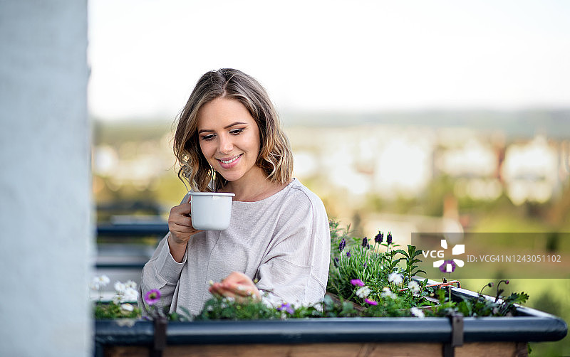 快乐的年轻女子在阳台上的肖像与开花植物，放松与咖啡。图片素材