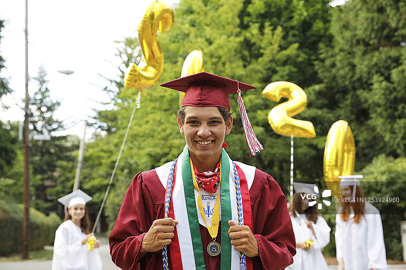 毕业典礼上的一群青少年，都戴着帽子，穿着长袍。他们站在附近的街道上，举着一个巨大的金色氦气球，上面写着2020年。镜头聚焦在前景中的一个对着镜头微笑的男孩身上。图片素材