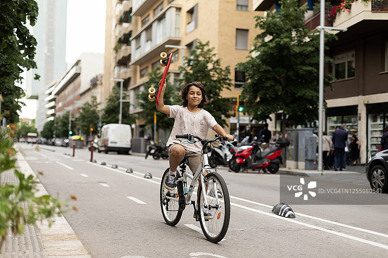 微笑的男孩拿着滑板，骑着自行车在街道上图片素材