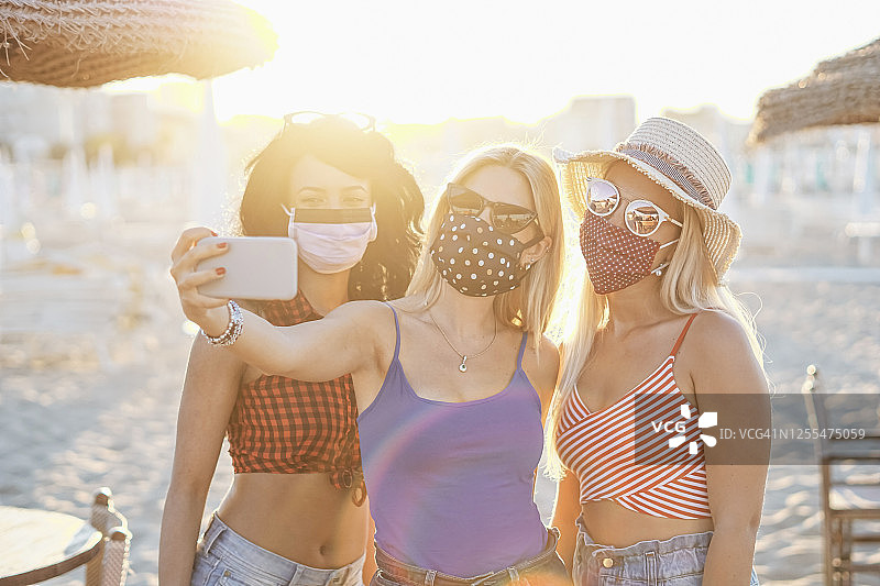 戴着口罩在户外自拍以保护冠状病毒的女孩——在海滩度假的青少年享受日落——多种族青年一起玩乐——使用智能手机的年轻人图片素材