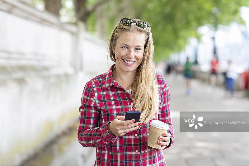 微笑的女人拿着咖啡用手机站在人行道上图片素材