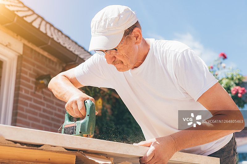 木匠老人用钻头在木板上钻孔或用电锯切割木头。木工，家装，自己动手(DIY)和建筑工程概念，行动阳光镜头。图片素材