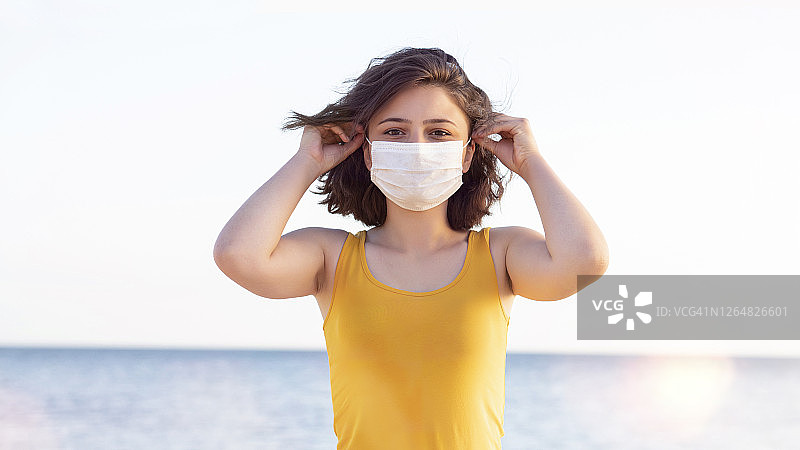 年轻的成年妇女戴着污染口罩以保护自己免受病毒感染。年轻女子戴上防护面具的肖像，夏天，假期，大海，海滩。图片素材