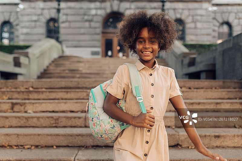 一个快乐的女孩站在学校大楼前的肖像图片素材