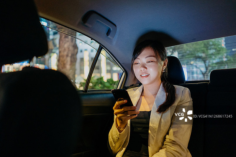 在一个夜晚的城市里，年轻的亚洲女商人坐在乘用车座位上使用智能手机图片素材