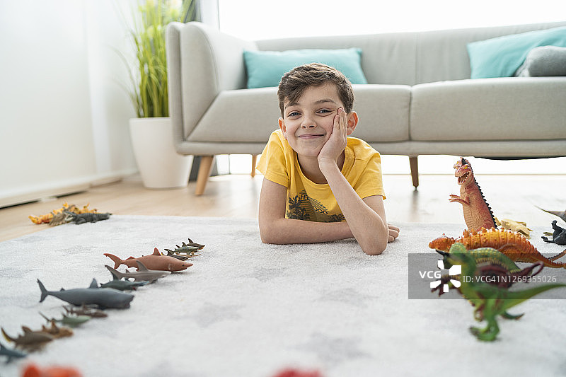 一个手托着下巴，面带微笑的男孩躺在客厅地毯上的玩具动物旁边图片素材