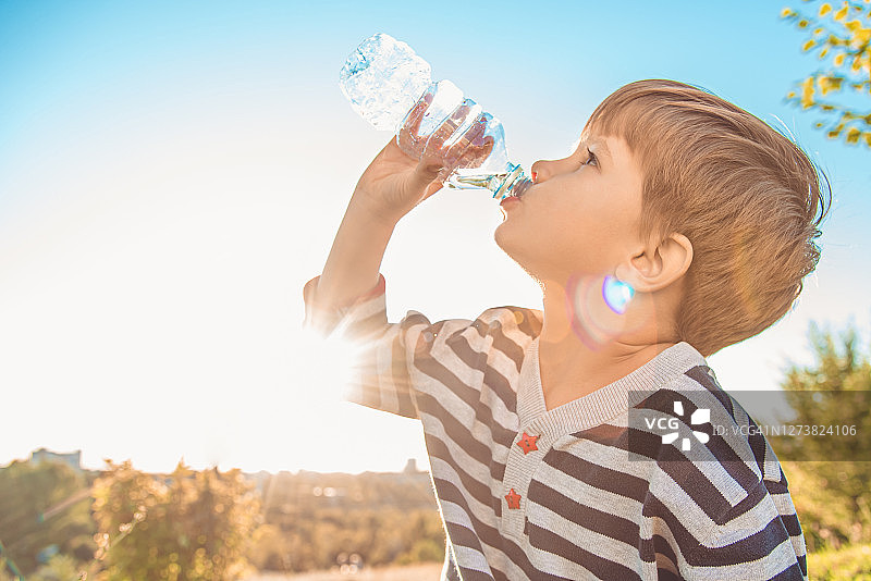 夏天日落时，一个漂亮的孩子坐在草地上喝瓶子里的水。男孩在热天解渴图片素材