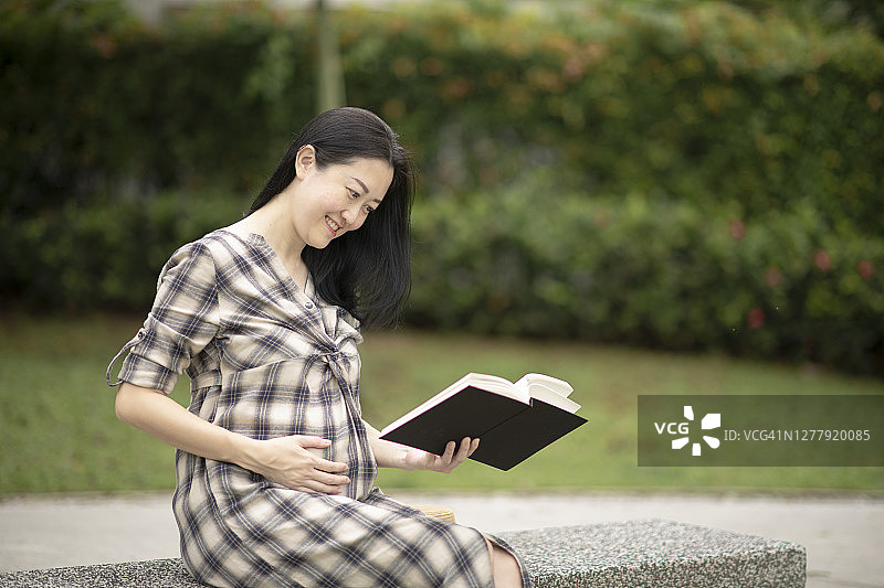 亚裔华人孕妇坐在公园长椅上看书图片素材
