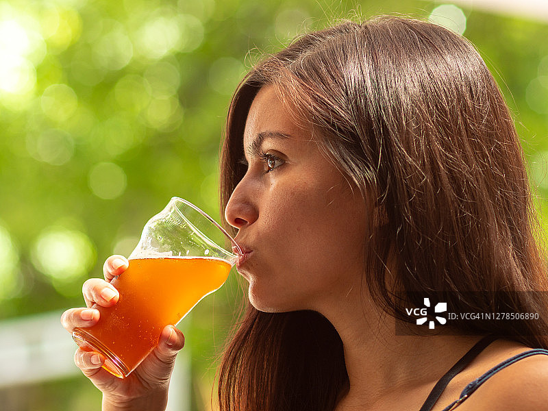 西班牙年轻女子在绿色树叶模糊的背景下，在大玻璃杯中喝红色手工啤酒图片素材