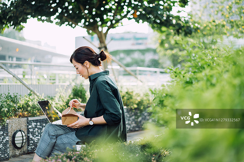 一位年轻的亚洲职业女商人坐在城市公园的长椅上，在午休时间用笔记本电脑工作，手里拿着一盒健康沙拉和一杯咖啡图片素材