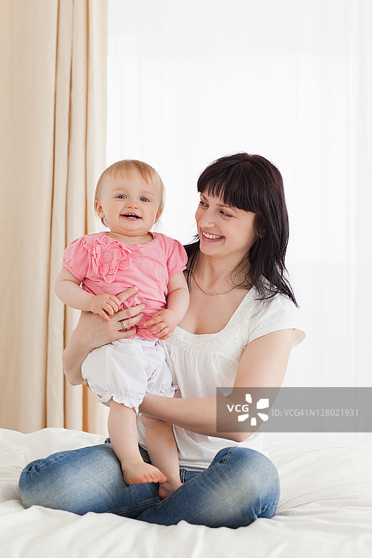 可爱的黑发女人抱着她的孩子在她的膝盖上，坐在床上图片素材