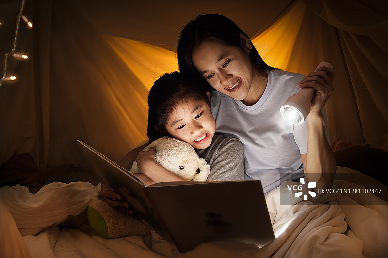 家庭的概念。母亲和孩子在睡觉前一起在儿童帐篷里用手电筒看书。快乐的妈妈在床单帐篷里给女儿读故事书图片素材