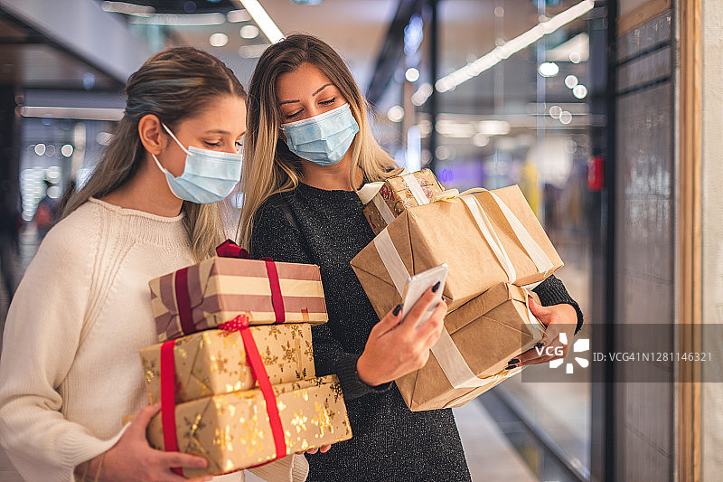 新冠肺炎疫情期间，两个美丽的女孩在购物中心用手机购买圣诞礼物。图片素材