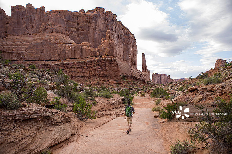 一名男子在被高高的红色岩石悬崖环绕的风景优美的小径上徒步。图片素材