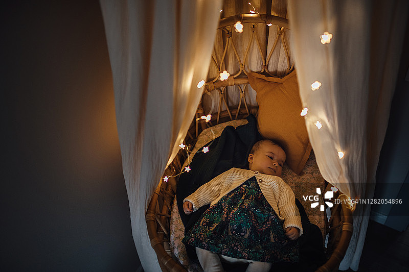 睡意朦胧的男孩在舒适的婴儿床里打盹，家里有顶棚和花环。高角度的婴儿睡觉与照明灯的藤条摩西篮子。圣诞装饰。图片素材