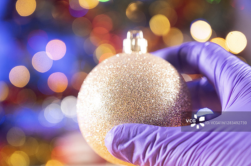 手持圣诞装饰球的人戴着防护手套。背景是一棵装饰过的圣诞树。图片素材
