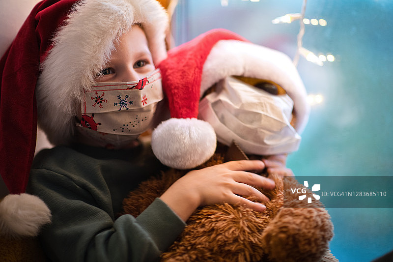 可爱的小男孩戴着防护面具和圣诞帽和他的泰迪熊在圣诞节图片素材