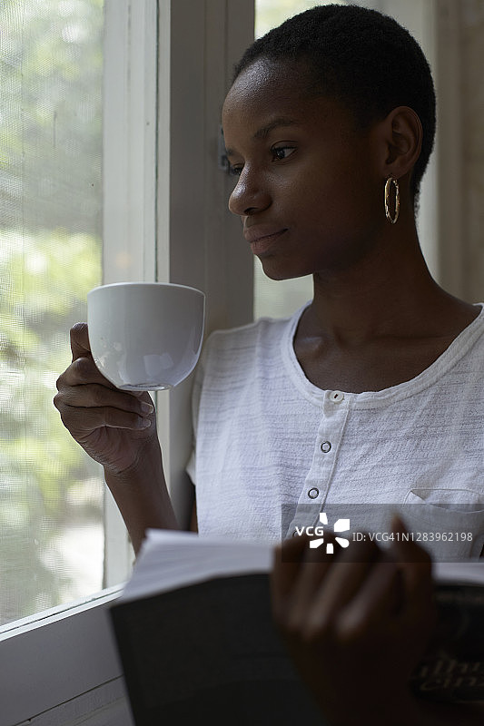 特写的体贴的年轻女子拿着咖啡和书，透过窗户看在家里图片素材