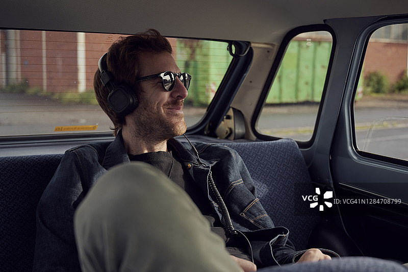 一个年轻人坐在汽车后座上，一边戴着耳机听音乐，一边四处张望。图片素材