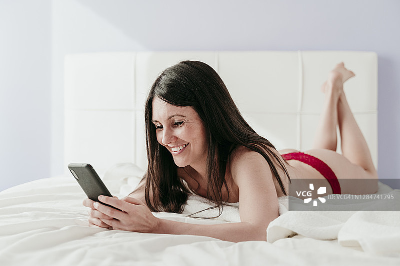 微笑美丽的女人使用智能手机，而躺在床上半穿衣在家图片素材