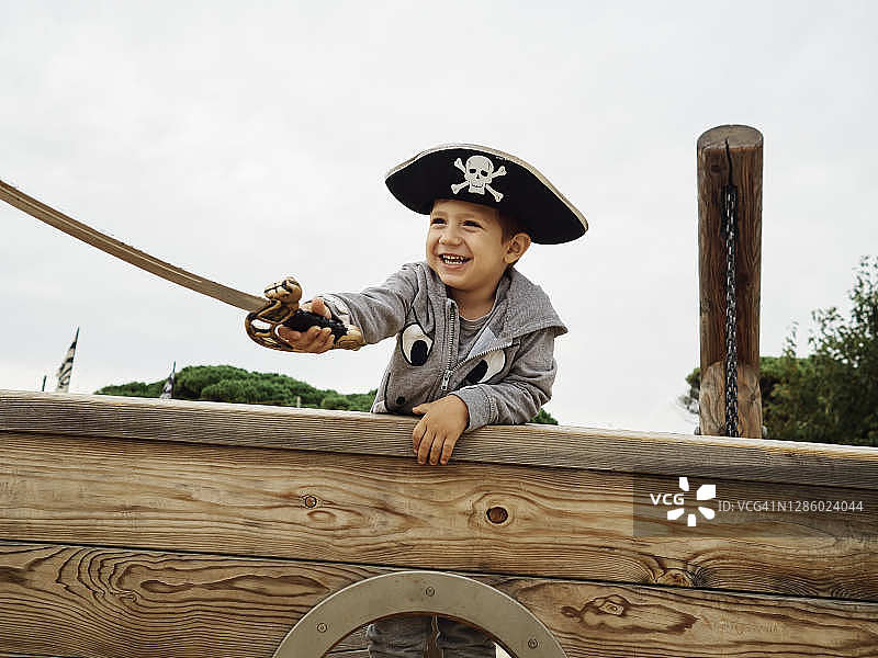 小男孩戴着海盗帽，在海盗船里微笑着，手里拿着一把剑图片素材