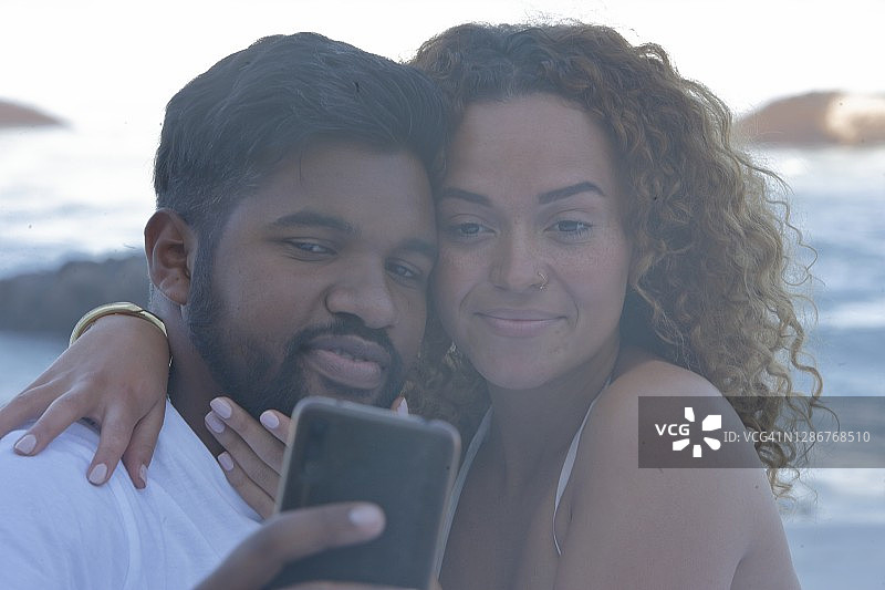 一对美丽的夫妇一边在海滩上自拍，一边用手机做鬼脸图片素材