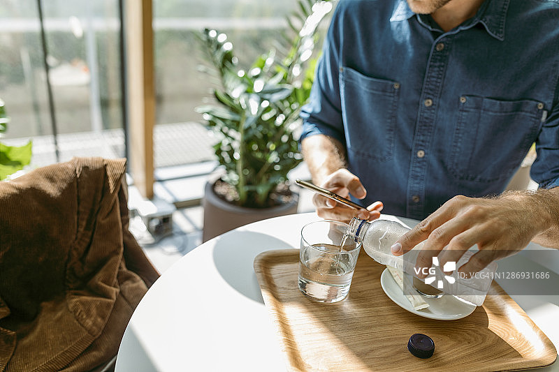 年轻的商人在咖啡馆的桌子上一边往玻璃杯里倒水一边用智能手机图片素材