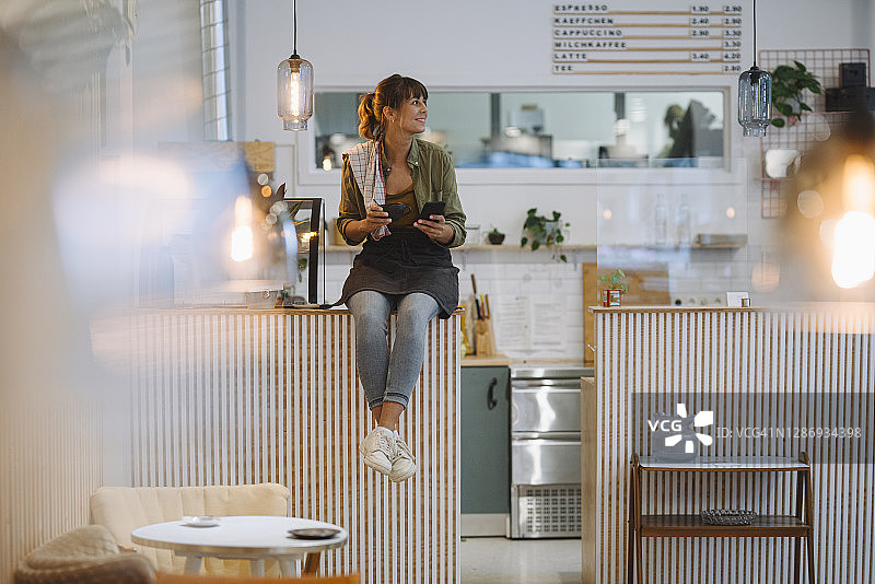年轻的女商人拿着咖啡看着别处，用智能手机坐在咖啡馆的柜台上图片素材