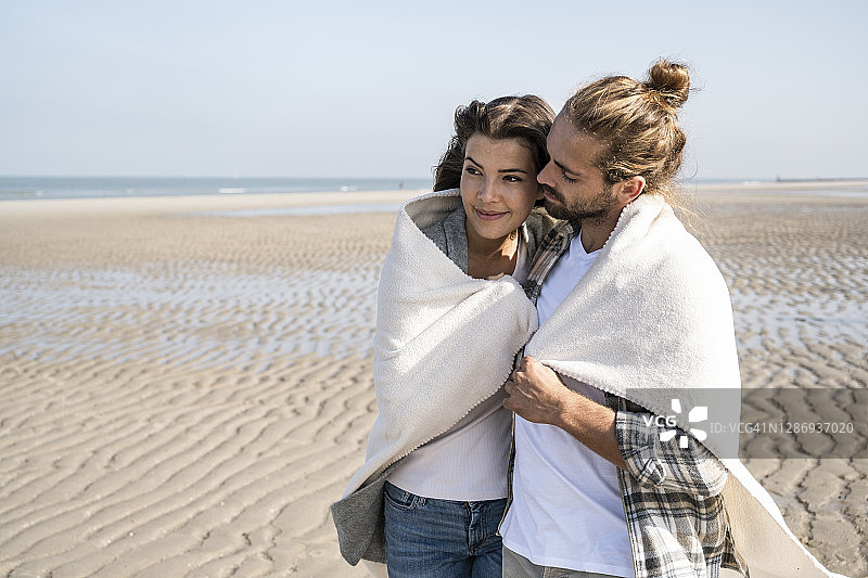 一对浪漫的年轻夫妇裹着毯子站在海滩上图片素材