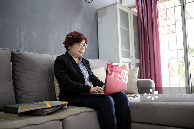 亚洲华裔商务女性坐在沙发上使用笔记本电脑在家工作图片素材