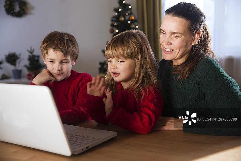 家庭母亲和两个孩子看着笔记本电脑，挥手，视频打电话给朋友或亲戚呆在家里过圣诞节。图片素材