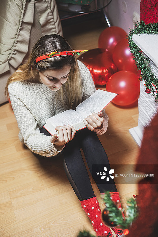 年轻女子在圣诞树下看书，在壁炉旁暖腿图片素材
