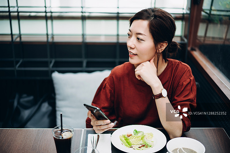 美丽年轻的亚洲女人坐在餐厅里吃健康的蔬菜沙拉作为午餐，手里拿着智能手机，眼睛看着别处，陷入沉思图片素材