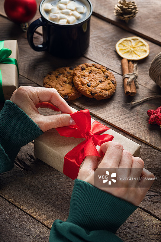 一个穿着绿色毛衣的女孩或女人拿着并系着圣诞礼物。准备一个家庭假日的概念，快乐的平安夜。木桌的背景上放着热咖啡、棉花糖、巧克力饼干。图片素材