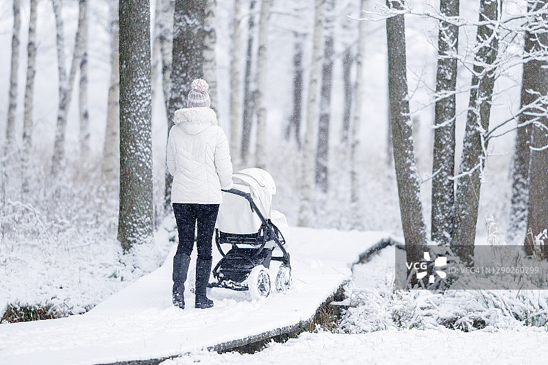 一名年轻的成年母亲推着婴儿车，走在白雪覆盖的木制小道上。新鲜的第一场雪。享受和平的漫步。后视图。图片素材