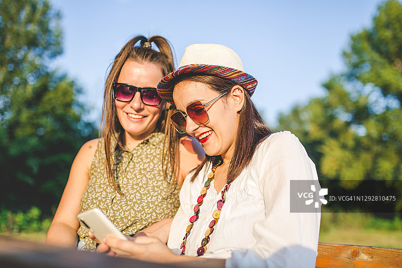 两个开心的女人在城市公园的长椅上玩智能手机图片素材