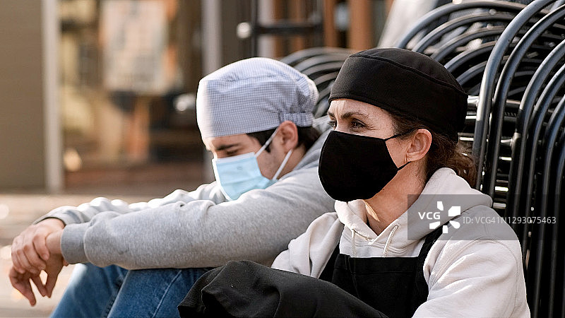 一名成熟的女厨师和一名戴着防护口罩的年轻男同事坐在他们工作的餐厅外面的地板上图片素材