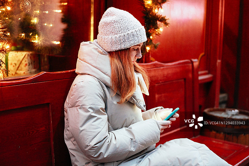 年轻快乐微笑的女人拿着电话红头发在户外的街道上穿着温暖的冬装，圣诞节日市场装饰着仙女灯和树在夜晚。生活方式度假。图片素材