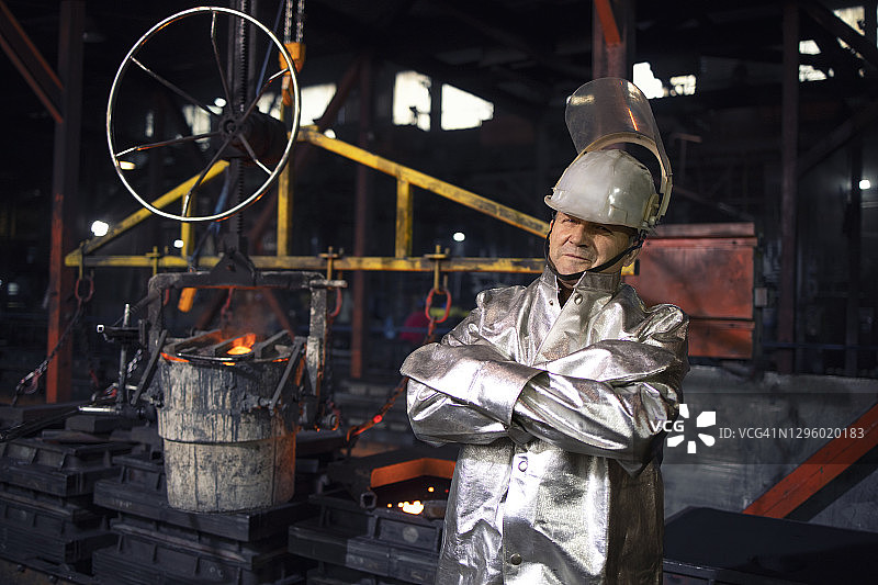 一名身穿铝化高温防护服的工人双臂交叉站在铸钢制造厂。背景桶内装有液态铁水。冶金和重工业。图片素材